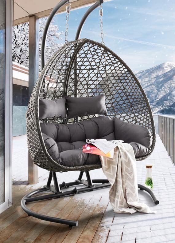 Großer grauer Hängesessel auf verschneiter Terrasse mit Kissen, Decke und Zeitschriften, Blick auf winterliche Berge