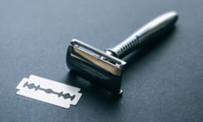 Ein klassischer Nassrasierer aus Metall liegt neben einer einzelnen Rasierklinge auf einer dunklen Oberfläche.