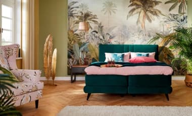 Ein stilvolles Schlafzimmer mit einem tiefgrünen Bett, das mit bunter Bettwäsche in Rosa- und Türkistönen dekoriert ist. Im Hintergrund ist eine tropische Wandtapete mit Pflanzenmotiven zu sehen. Ein Sessel mit floralem Muster ergänzt die Einrichtung.