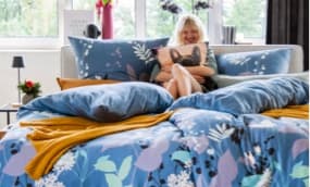 Ein gemütliches Schlafzimmer mit einem großen Bett, das mit bunten, floral gemusterten Bettwäsche und Kissen dekoriert ist. Auf dem Bett liegen auch eine gelbe Decke und ein Kissen mit einem Hundemotiv. Im Hintergrund sind Fenster mit Blick ins Freie und
