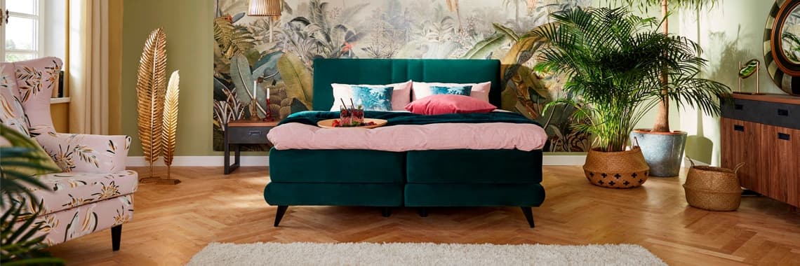 Ein stilvolles Schlafzimmer mit einem tiefgrünen Bett, das mit bunter Bettwäsche in Rosa- und Türkistönen dekoriert ist. Im Hintergrund ist eine tropische Wandtapete mit Pflanzenmotiven zu sehen. Ein Sessel mit floralem Muster ergänzt die Einrichtung.