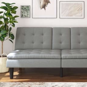Ein modernes, graues Sofa mit einer gepolsterten Oberfläche und Knopfdetails. Im Hintergrund hängen drei Bilder an der Wand, und eine große Pflanze steht links neben dem Sofa. Der Boden ist aus Holz und ein dekorativer Teppich liegt darunter.