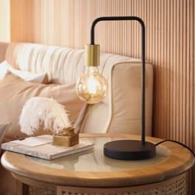 Eine stilvolle Tischlampe mit einem schwarzen Gestell und einer großen, gläsernen Glühbirne, die warmes Licht ausstrahlt. Neben der Lampe liegt ein Buch auf einem runden, hellen Tisch, Die Wand hat eine strukturierte Holzverkleidung.