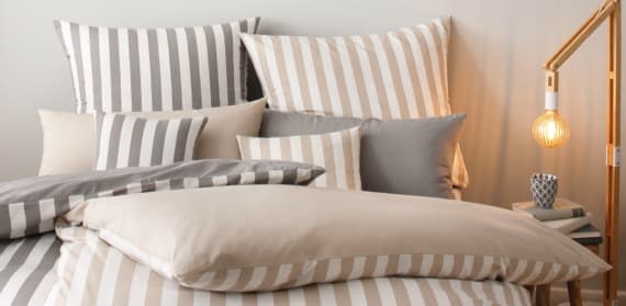Ein einladendes Bett mit gestreifter Bettwäsche in sanften Beige- und Grautönen. Verschiedene Kissen in passenden Farben sind dekorativ angeordnet. Neben dem Bett steht eine stilvolle Lampe mit einem warmen Licht, die eine gemütliche Atmosphäre schafft.