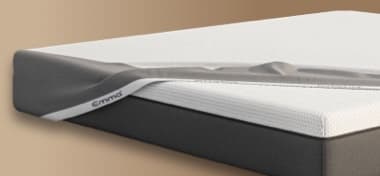 Eine Matratze mit einem abnehmbaren Bezug, der teilweise heruntergezogen ist, um die darunterliegende Struktur zu zeigen. Die Matratze hat eine graue Unterseite und eine weiße, atmungsaktive Oberseite. Der Hintergrund ist neutral und hell gehalten.