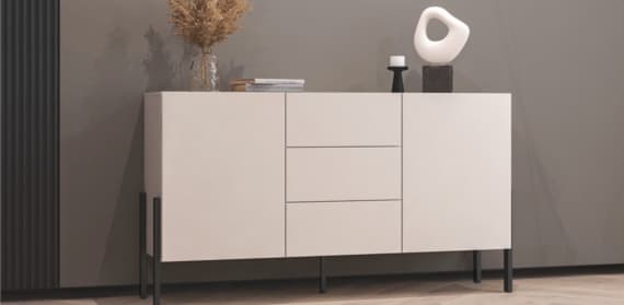 Ein modernes Sideboard in hellem Farbton mit zwei Türen und drei Schubladen. Auf dem Sideboard stehen eine Vase mit Trockenblumen, eine Kerze und eine dekorative Skulptur. Der Hintergrund ist in einem neutralen Farbton gehalten, der Boden ist aus Holz.