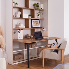 Eine moderne Schreibtisch-Regal-Kombination, auf dem ein Laptop und eine Schreibtischlampe stehen. Neben der Schreibtisch-Kombi steht ein bequemer Stuhl mit einem bunten Kissen. Der Boden ist aus Holz, und die Atmosphäre ist hell und einladend.