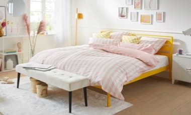 Ein helles und einladendes Schlafzimmer mit einem gelben Bett, das mit gestreifter Bettwäsche in sanften Rosa- und Weißtönen dekoriert ist. Vor dem Bett steht eine elegante Bank, und im Hintergrund sind Regale mit dekorativen Objekten.