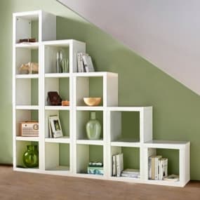 Ein modernes Würfel-Regal in Weiß, das an einer schrägen Wand steht. Es hat verschiedene Fächer, die mit Büchern, Pflanzen und dekorativen Objekten gefüllt sind. Die Wand ist in einem sanften Grünton gestrichen, und der Boden ist aus Holz, was