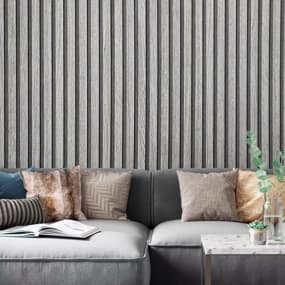 Eine gemütliche Couch in Grau mit mehreren dekorativen Kissen in verschiedenen Farben und Mustern. Im Hintergrund ist eine strukturierte Wand mit vertikal gestreifter Vliestapete. Neben der Couch steht ein kleiner Tisch mit einer Pflanze und einem Buch.