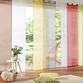 Ein heller Raum mit großen Fenstern, die mit transparenten Vorhängen in Gelb, Rot, Weiß, Braun und Grau dekoriert sind. Vor den Fenstern steht ein kleiner Tisch mit einer Pflanze. Auf dem Boden liegen ein weicher Teppich und 2 Kissen.