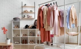 Ein stilvoller Raum mit einem Kleiderständer, an dem verschiedene Kleidungsstücke in sanften Farben hängen. Im Hintergrund sind Regale mit Schuhen, Taschen und Accessoires. Einladende Atmosphäre. Die Wände sind in einem hellen Farbton gehalten.