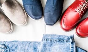 Eine Anordnung von verschiedenen Schuhen auf einem weißen Hintergrund. Zu sehen sind graue und blaue Schuhe sowie rote Schnürschuhe. Darunter liegt eine Jeansjacke, die dem Bild einen lässigen Look verleiht.