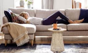 Eine Person liegt auf einem hellen Sofa, umgeben von Kissen und einer Decke. Neben dem Sofa steht ein kleiner Tisch. Im Hintergrund sind Fenster, die viel Tageslicht hereinlassen für eine gemütliche Atmosphäre.