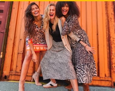 Drei Frauen in gemusterten Kleidern posieren vor einer orangefarbenen Wand.