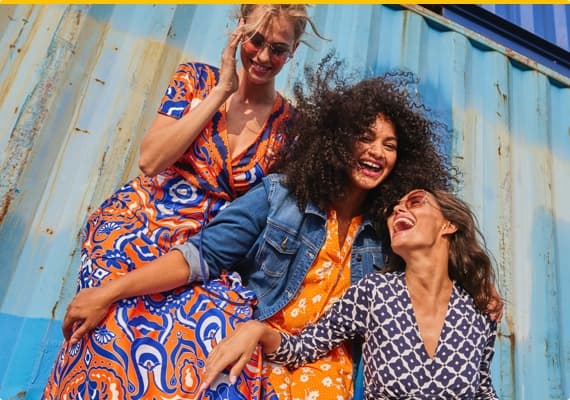 Drei Frauen in farbenfrohen Kleidern, eine trägt Jeansjacke, vor einem Transport-Container