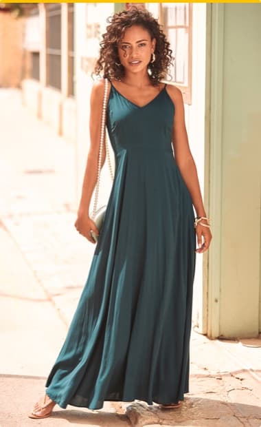 Frau in langem, ärmellosem, dunkelgrünem Kleid mit V-Ausschnitt steht auf Gehweg und hält kleine Tasche