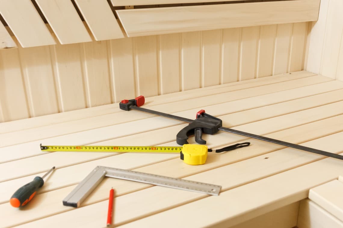 Werkzeuge auf einer hellen Holzoberfläche in einer Sauna: Maßband, Winkel, Schraubenzwinge, Schraubenzieher und ein roter Stift.