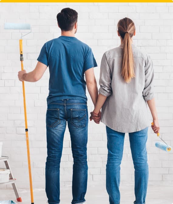 Paar steht von hinten vor weißer Wand, Mann hält Farbroller, Frau hält kleineren Farbroller, beide tragen Jeans