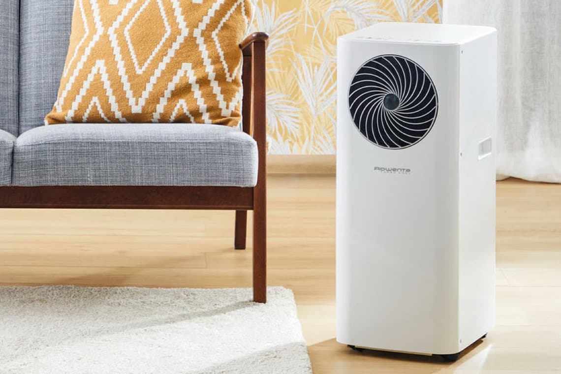 Ein weißes Klimagerät steht neben einem grauen Sofa mit einem orangefarbenen Kissen. Der Boden ist aus Holz, und ein Teppich liegt davor.