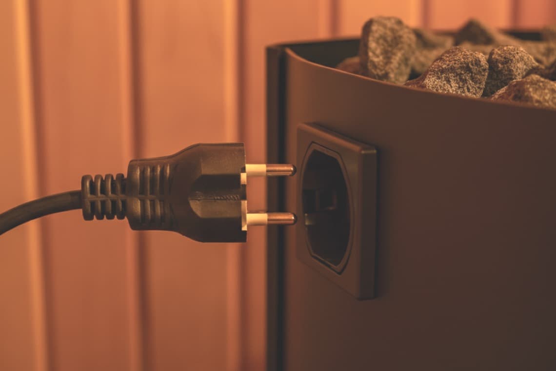 Ein schwarzes Stromkabel ist an eine Steckdose neben einem Saunaofen angeschlossen. Im Hintergrund sind Steine im Ofen sichtbar, Holzverkleidung umgibt die Szene.