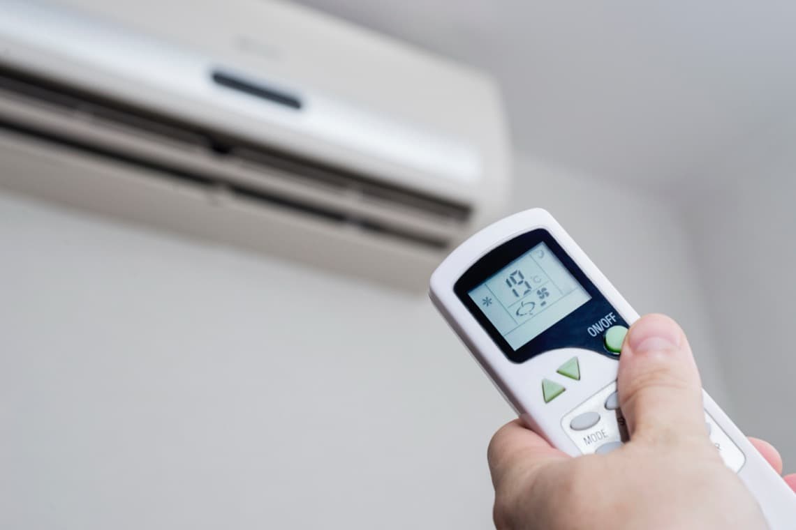 Hand hält eine Fernbedienung mit digitalem Display mit der eine Klimaanlage an der Wand gesteuert wird