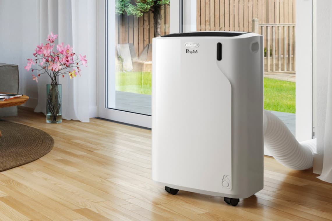 Ein weißes Klimagerät steht auf einem hellen Holzfußboden neben einem Fenster. Draußen ist ein grüner Garten sichtbar. Eine Vase mit rosa Blumen steht daneben.