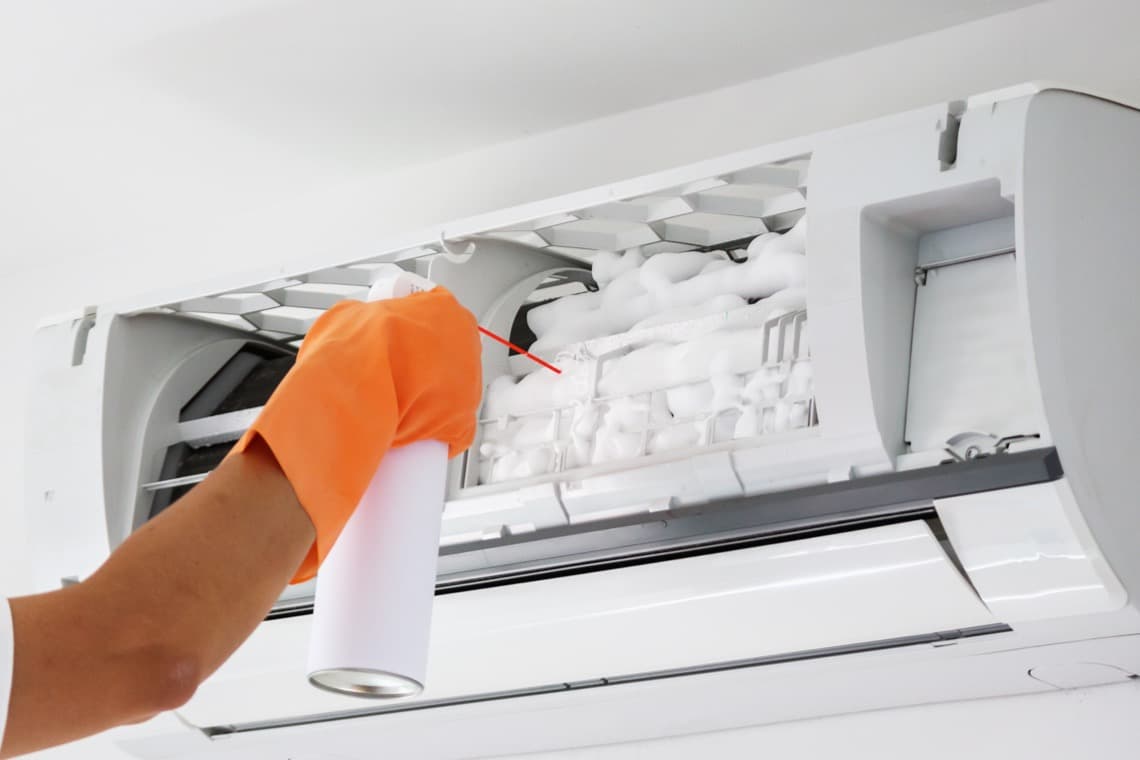 Eine Hand mit einem orangefarbenen Handschuh sprüht Kältemittel in eine Wand-Klimaanlage, die geöffnet ist. Der Hintergrund ist hell.