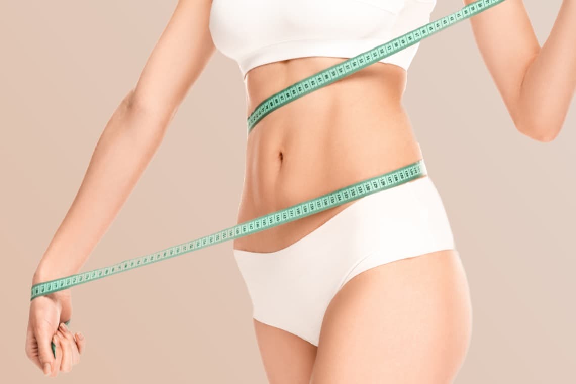 Eine Person in einem weißen Oberteil und Unterwäsche hält ein grünes Maßband um die Taille. Der Hintergrund ist hell und neutral.