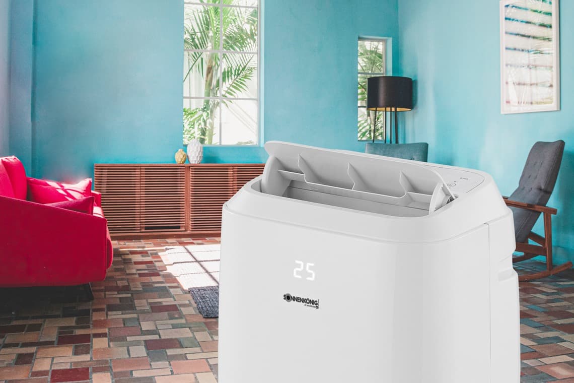 Ein weißes Klimagerät steht in einem hellen Raum mit blauen Wänden. Im Hintergrund sind ein rotes Sofa und ein Tisch mit Pflanzen zu sehen.