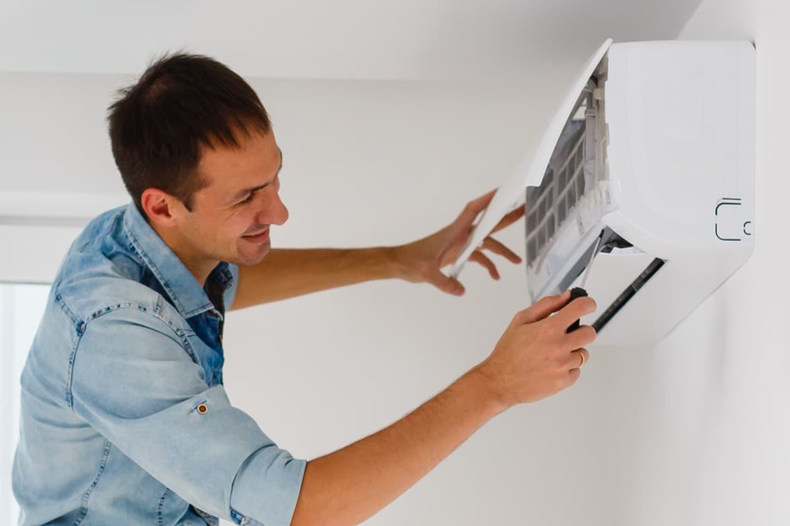 Eine Person in einem blauen Hemd wartet an einer Wand-Klimaanlage, während sie die Abdeckung öffnet, um Wartungsarbeiten durchzuführen.