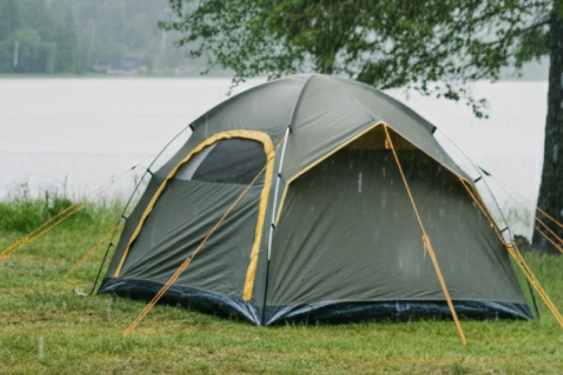 Ein grünes Zelt steht im Regen auf einer Wiese, nahe einem See. Bäume sind im Hintergrund sichtbar.
