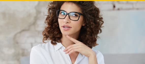 Frau mit lockigem Haar in einem weißen Hemd, nachdenklich mit der Hand am Kinn, vor einer hellen Wand.