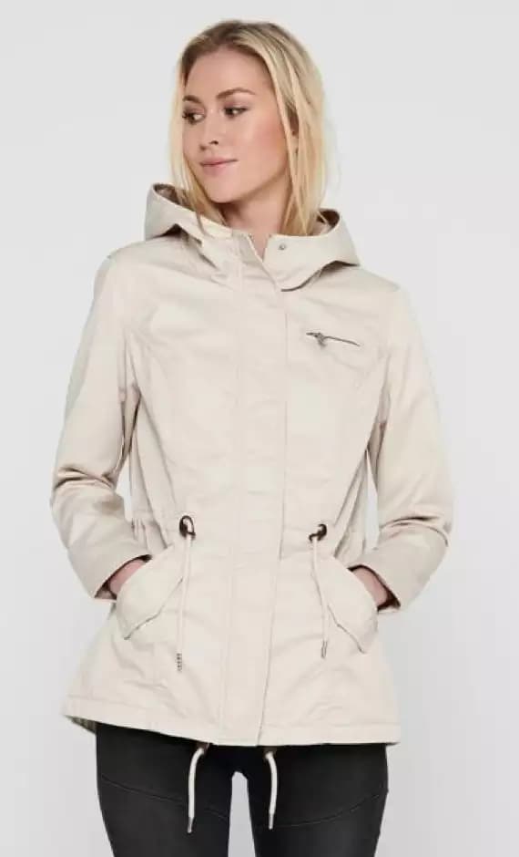 Frau trägt beige Jacke mit Kapuze und Taschen, Gesicht ist verschwommen, schwarze Hose.