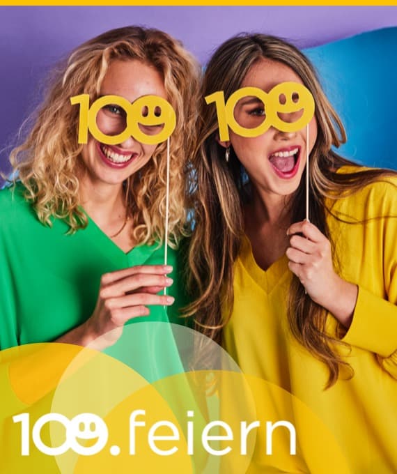 100 feiern – Zwei Personen in grünen und gelben Shirts halten Schilder mit der Zahl 100 vor buntem Hintergrund