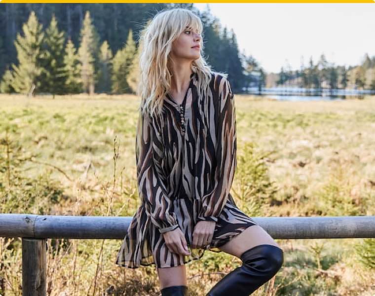 Frau sitzt im Schneidersitz auf einem Spiegel im Freien, trägt bunte Bluse mit Blumenmuster und blaue Jeans