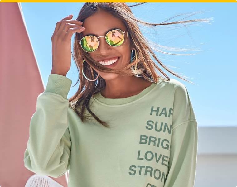 Frau in grünem Pullover mit Schriftzug, hebt Hand an die Stirn, die Haare wehen im Wind.