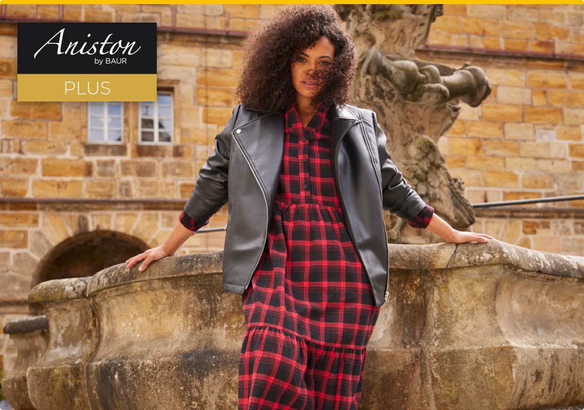 Aniston by BAUR PLUS – Frau in rotem Karokleid und schwarzer Lederjacke lehnt an einem Brunnen vor historischer Gebäude.