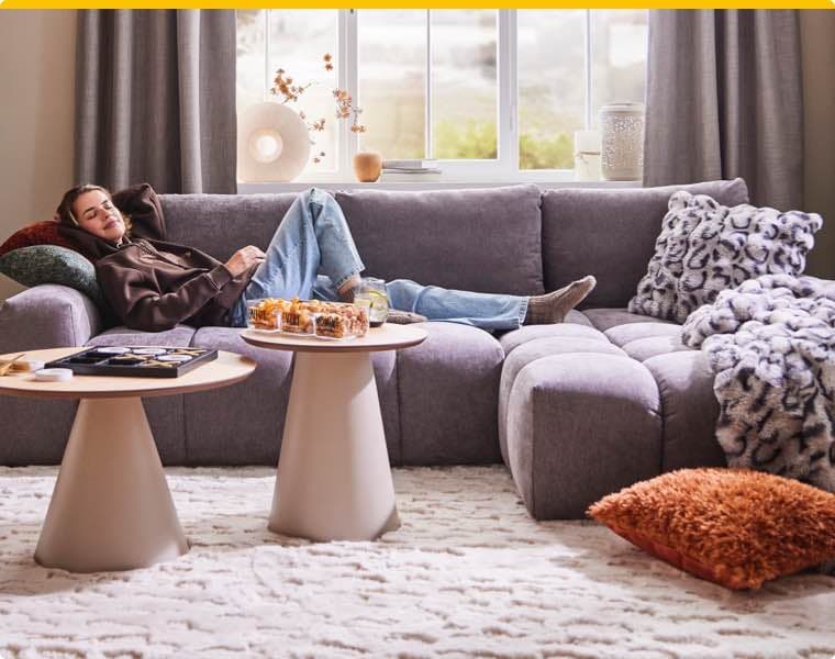 Person liegt entspannt auf grauem Sofa, Snacks und Getränke auf zwei runden Tischen, gemütliche Kissen und Fenster im Hintergrund
