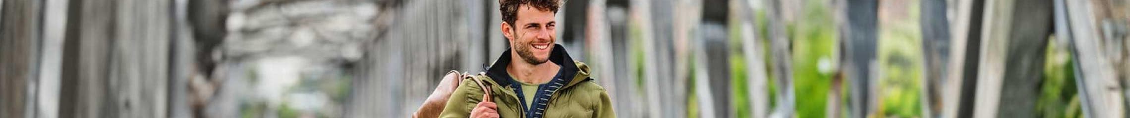 Person mit beigem Rucksack und olivgrüner Jacke geht auf einem Weg mit Bäumen und Brücken im Hintergrund.