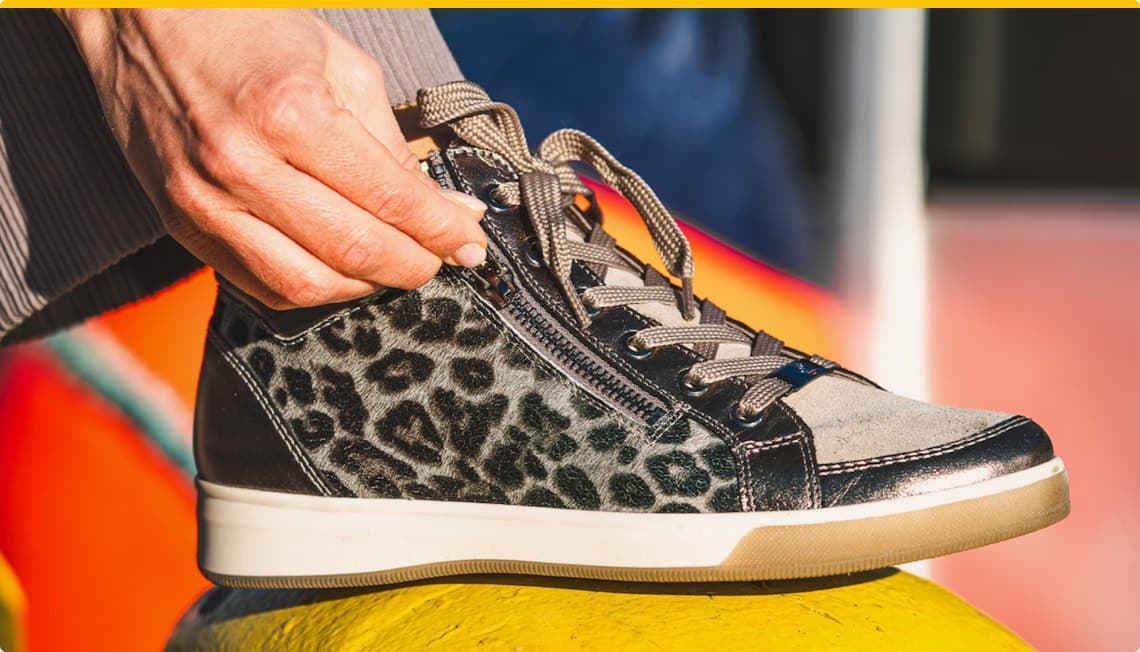 Hand zieht Reißverschluss an einem grau-schwarzen Sneaker mit Leopardenmuster zu, im Hintergrund bunte Farben
