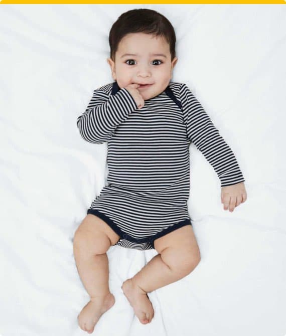 Baby in gestreiftem Einteiler liegt auf weißem Bett und hält eine Hand vor das Gesicht.