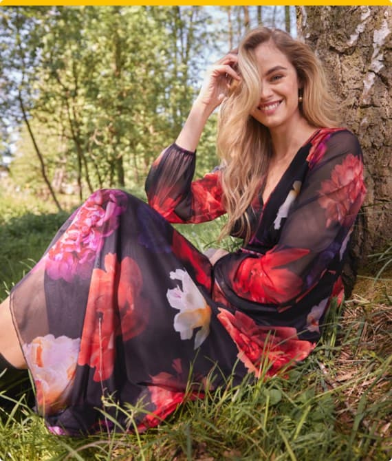 Frau mit langen Haaren sitzt im Wald und trägt ein langes Kleid mit buntem Blumenmuster