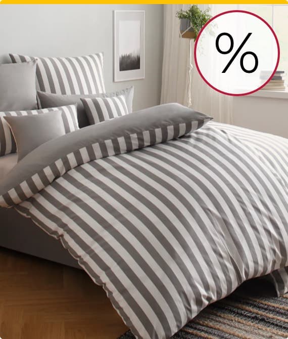 % – Grau-weiß gestreifte Bettwäsche auf einem Bett mit mehreren Kissen im gleichen Design. Fenster und Pflanzen im Hintergrund.