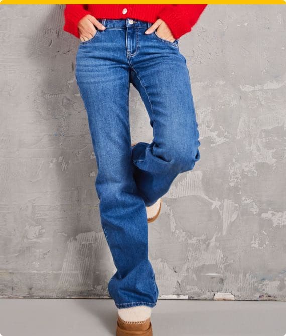 Person trägt blaue Jeans, rotes Oberteil und braune Stiefel, steht vor einer grauen Wand mit einer Hand in der Tasche
