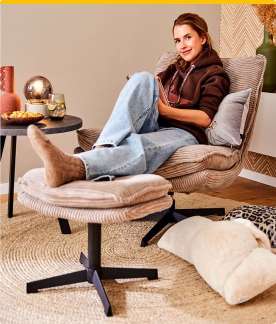Person sitzt entspannt in einem beigen Sessel, mit Füßen auf Hocker, trägt braunen Hoodie und Jeans, neben einem Tisch mit Snacks.