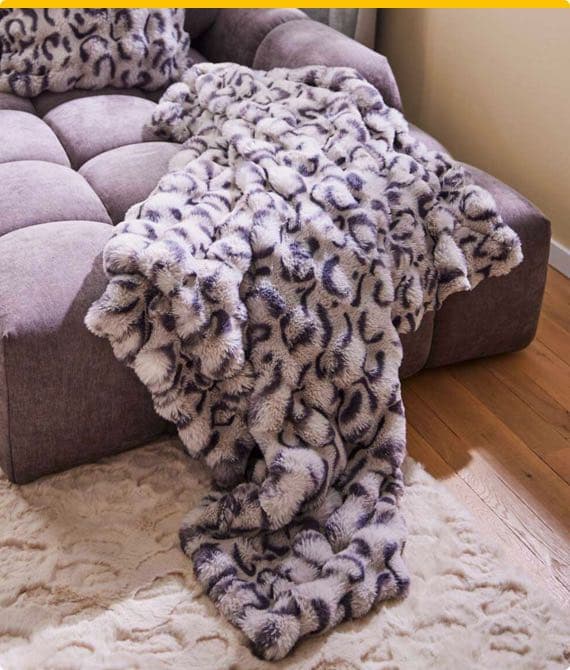 Weiche, gemusterte Decke über lila Sofa drapiert, auf hellem Teppichboden im Wohnzimmer.