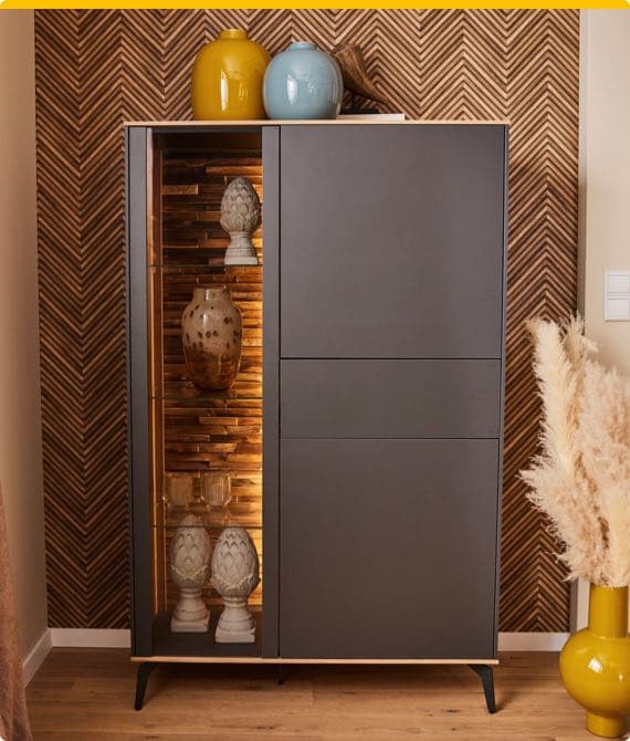 Dunkler Schrank mit dekorativen Vasen und Skulpturen in beleuchtetem Regal. Gelbe und blaue Vasen oben, Pampasgras daneben.