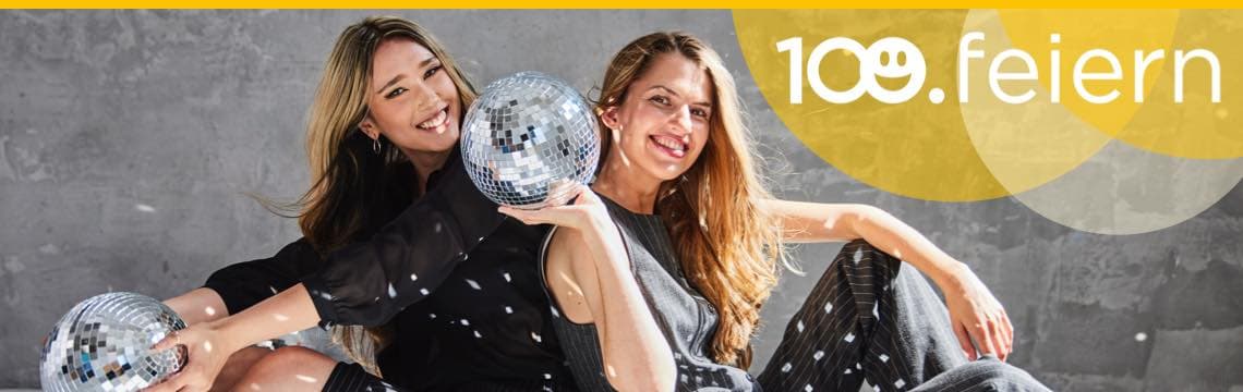 100 feiern – Zwei Frauen sitzen mit Discokugeln am Boden, sie tragen festliche Kleidung und lachen