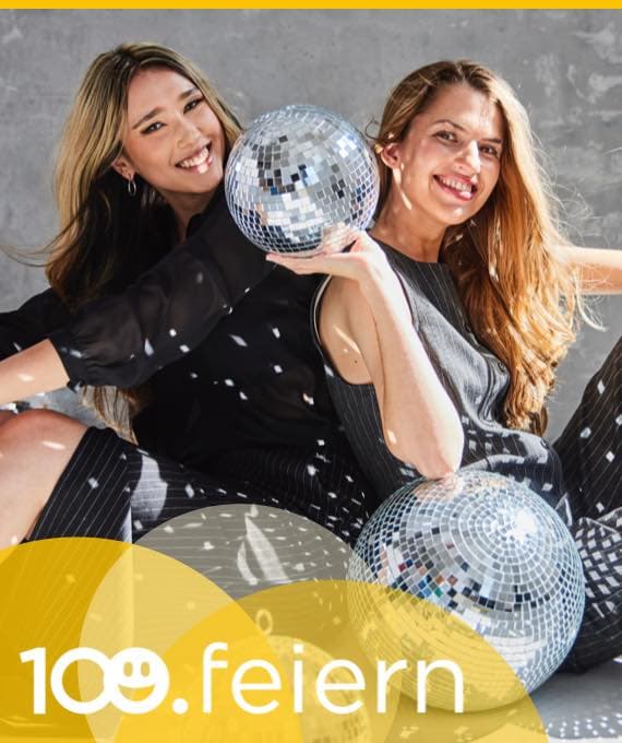 100 feiern – Zwei Frauen sitzen mit Discokugeln am Boden, sie tragen festliche Kleidung und lachen